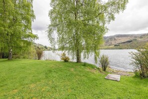 A grassy area with a wooden deck by a lake, surrounded by trees and hills in the background.