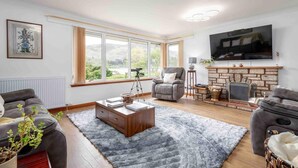 A living room with a fireplace, a large TV, and a window with a view of the outdoors. - 3 Bedroom Bungalow With Htub & Private Loch Access (Crieff)