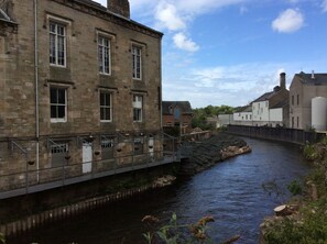 A stone building with multiple windows, a bridge over a river, and a white building in the background. - Lake District Western 2-bed Cabin, Pet Frien (Wigton)
