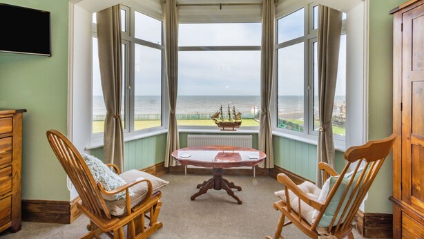 A room with a bay window overlooking the sea, featuring two rocking chairs, a small table, and a ship model. - Beachside Apartment (Hornsea)
