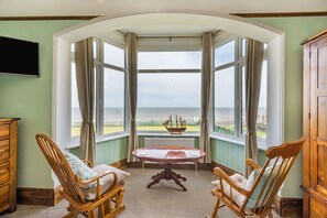 A room with a bay window overlooking the sea, featuring two rocking chairs, a small table, and a ship model.
