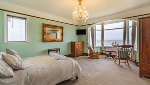 A bedroom with a large bed, a wooden dresser, a rocking chair, a table, and a view of the ocean through a large window. - Beachside Apartment (Hornsea)