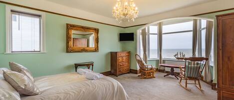 A bedroom with a large bed, a wooden dresser, a rocking chair, a table, and a view of the ocean through a large window.