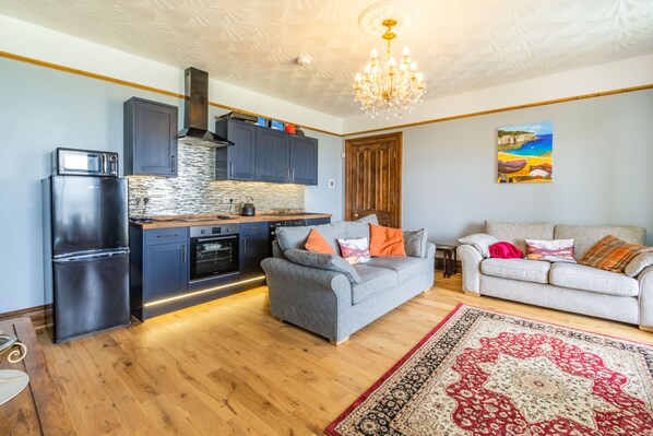 A modern kitchen with a built-in oven, a microwave, and dark cabinets. There is a grey sofa with orange and white cushions, and a red and beige patterned rug on the wooden floor.