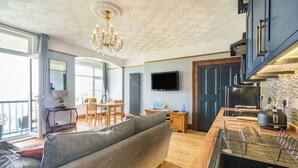 A modern living room with a grey sofa, wooden flooring, a chandelier, and a kitchen area with a black range hood and a TV mounted on the wall. - Beachside Apartment (Hornsea)