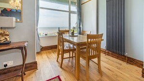 A dining area with a wooden table and chairs, a view of the sea through large windows, and a radiator on the wall. - Beachside Apartment (Hornsea)