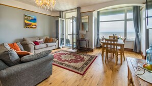 A living room with a sofa, a dining table with chairs, a chandelier, and a view of the sea through large windows. - Beachside Apartment (Hornsea)