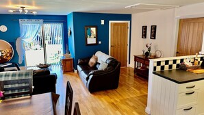 A living room with a black leather sofa, wooden dining table, and a kitchen area with a checkered backsplash. - Yr Encil (Gaerwen)