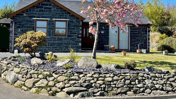 A stone house with a gabled roof, two visible windows, and a well-maintained garden with a rock border and flowering tree. - Yr Encil (Gaerwen)