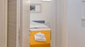 Un couloir étroit mène à une pièce où se trouvent un lit et une table jaune sur laquelle sont pliées des serviettes.