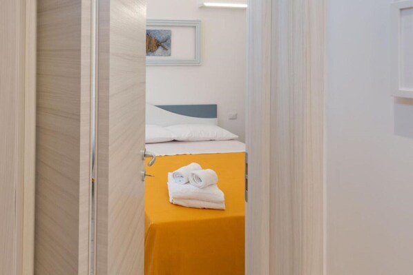 A narrow hallway leading to a room with a bed and a yellow table with folded towels.