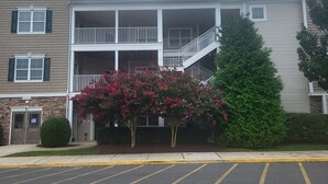 Ein mehrstöckiges Gebäude mit einem Balkon im zweiten Stock, einem Parkplatz davor und einem Baum mit rosa Blüten.