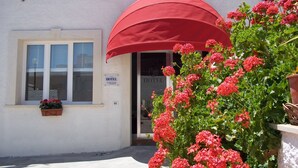 A hotel entrance with a red awning, a window with a potted plant, and a sign indicating the hotel's name and contact information. - Hotel Noto Marina a Mare Spa (Noto)