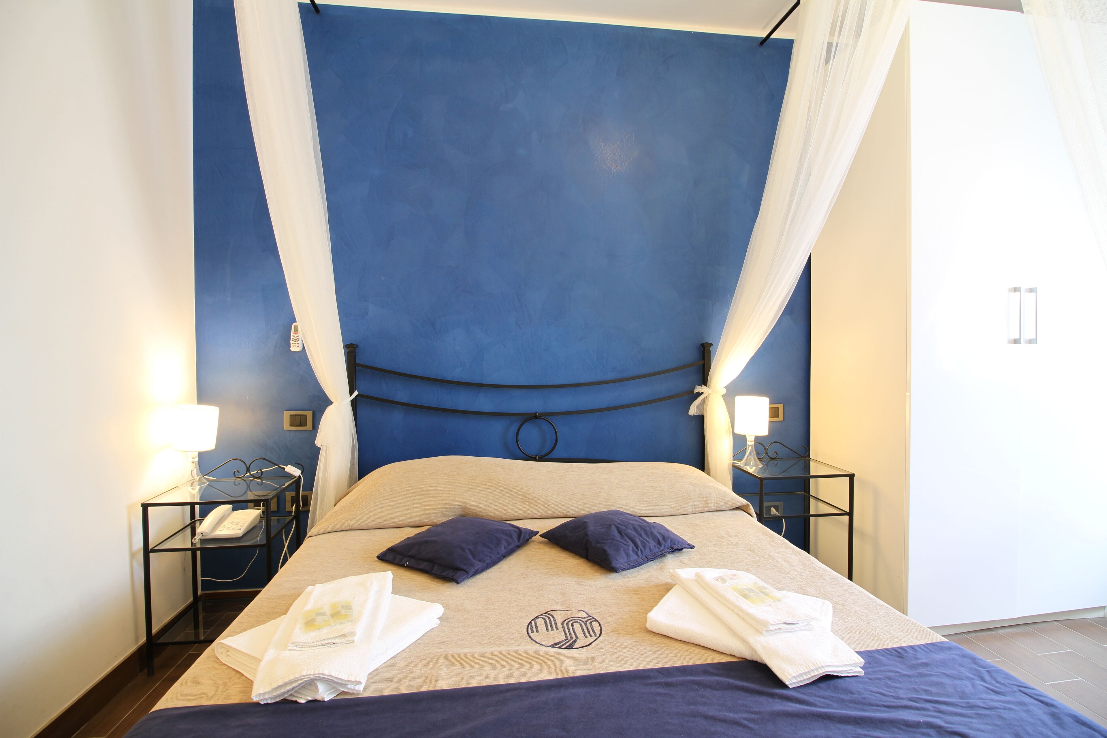 A neatly made bed with a blue headboard, two pillows, and a blanket with a logo. There are two bedside tables with lamps and a phone.