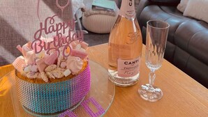 Ein gemütliches Wohnzimmer mit einem Glastisch, einer Flasche Canti Prosecco, einem Glas und einer Geburtstagstorte als Mittelpunkt.