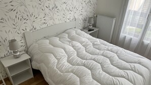 Ein ordentlich bezogenes Bett mit weißer Daunendecke, ein Nachttisch mit Lampe und eine Heizung.