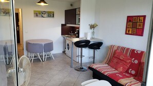 A compact living space with a kitchenette, a dining area with a round table and chairs, a sofa with floral upholstery, and a wall-mounted artwork. - Studio Mas du Tanit (Antibes)