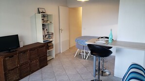 A compact living space with a television, a bookshelf, a dining area with a table and chairs, and a tiled floor. - Studio Mas du Tanit (Antibes)