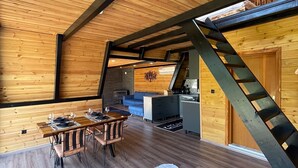 Een kamer in zolderstijl met houten wanden en een schuin dak. Er is een eethoek met een tafel en stoelen, een keuken met moderne apparatuur, en een gezellige zithoek met een bank en een salontafel.