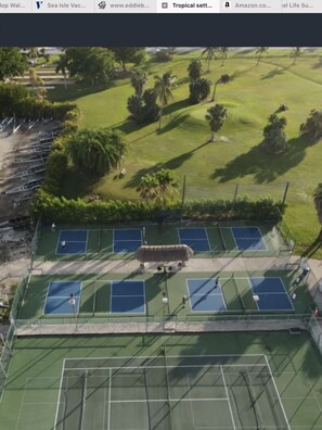 An aerial view of a tennis court complex with multiple courts, surrounded by greenery and palm trees.
