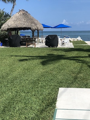 A grassy area with a thatched-roof structure, covered seating, and a view of the ocean.