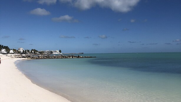 Une plage avec une eau turquoise et limpide, du sable blanc et quelques bâtiments en arrière-plan.