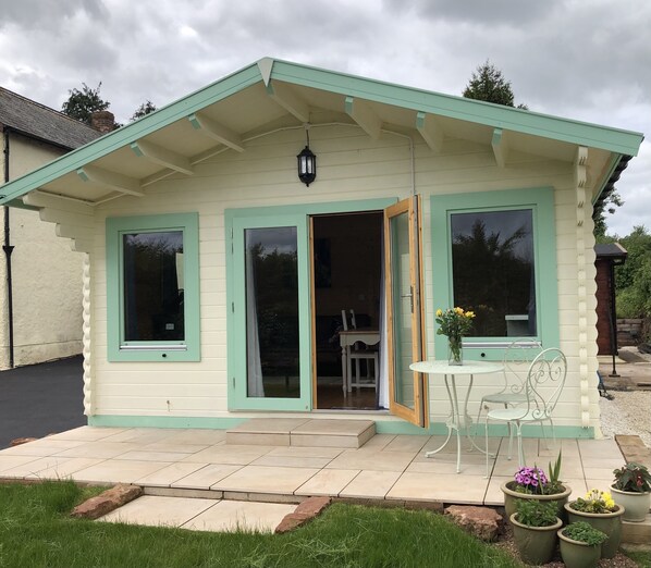 Une petite maison de campagne aux murs d’un vert pâle, dotée d’une terrasse carrelée, d’une table avec des chaises et de plantes en pot.
