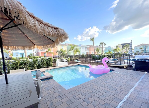 A swimming pool with a pink flamingo float, a thatched umbrella, and a brick deck.