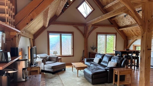 A spacious loft-style living room with a high ceiling, wooden beams, a fireplace, and large windows. - Post and Beam Carriage House in the heart of Mad River Valley (Waitsfield)