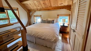 A cozy bedroom with a large bed, wooden ceiling beams, and a window overlooking greenery. - Post and Beam Carriage House in the heart of Mad River Valley (Waitsfield)
