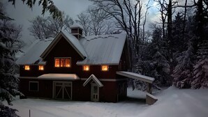 A snow-covered house with a garage, a chimney, and a porch. - Post and Beam Carriage House in the heart of Mad River Valley (Waitsfield)