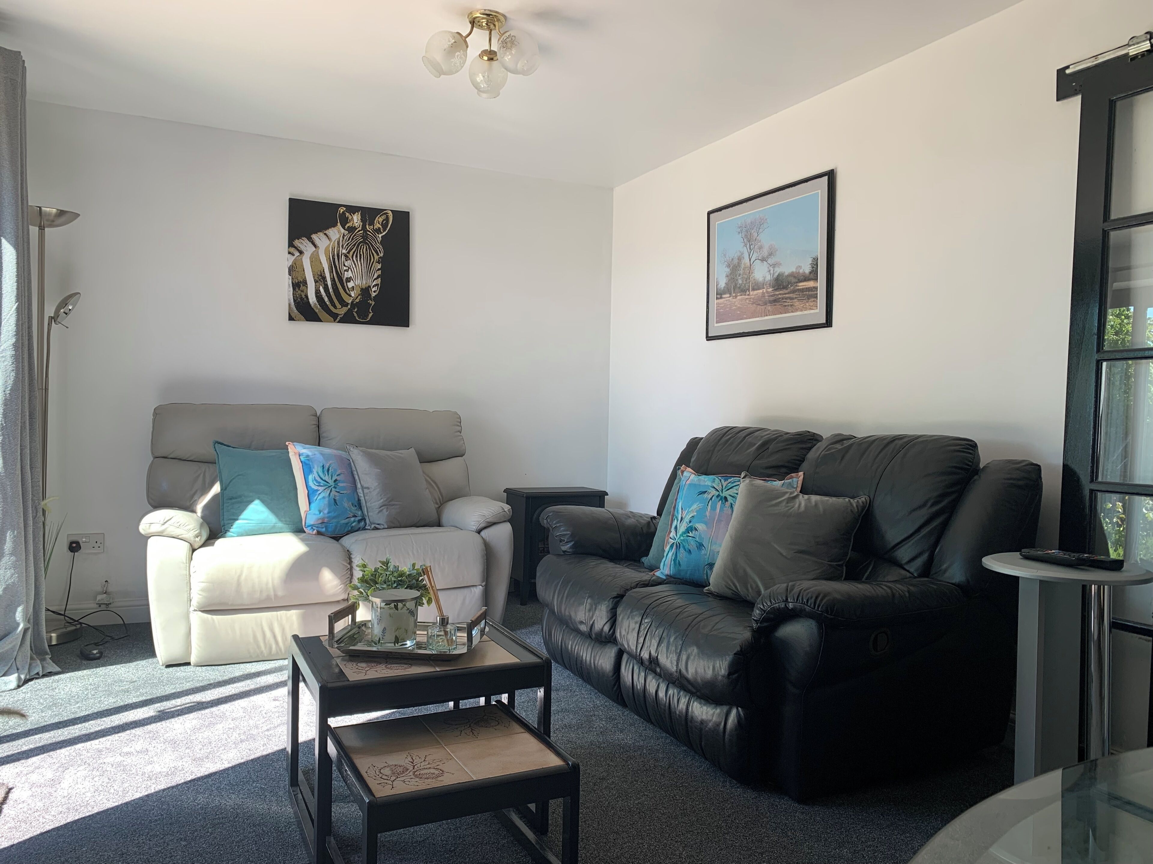 A living room with a beige sofa, a black leather sofa, and a coffee table. There are two wall-mounted pictures, one of a zebra and one of a landscape.