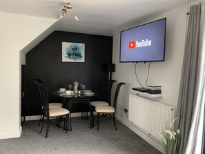 A dining area with a table set for four, a black-framed TV mounted on the wall, and a radiator below.