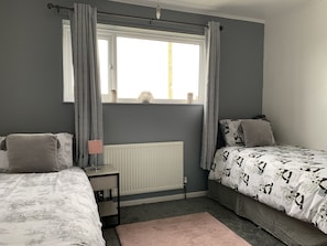 A bedroom with two beds, a window with curtains, a radiator, and a bedside table with a lamp.