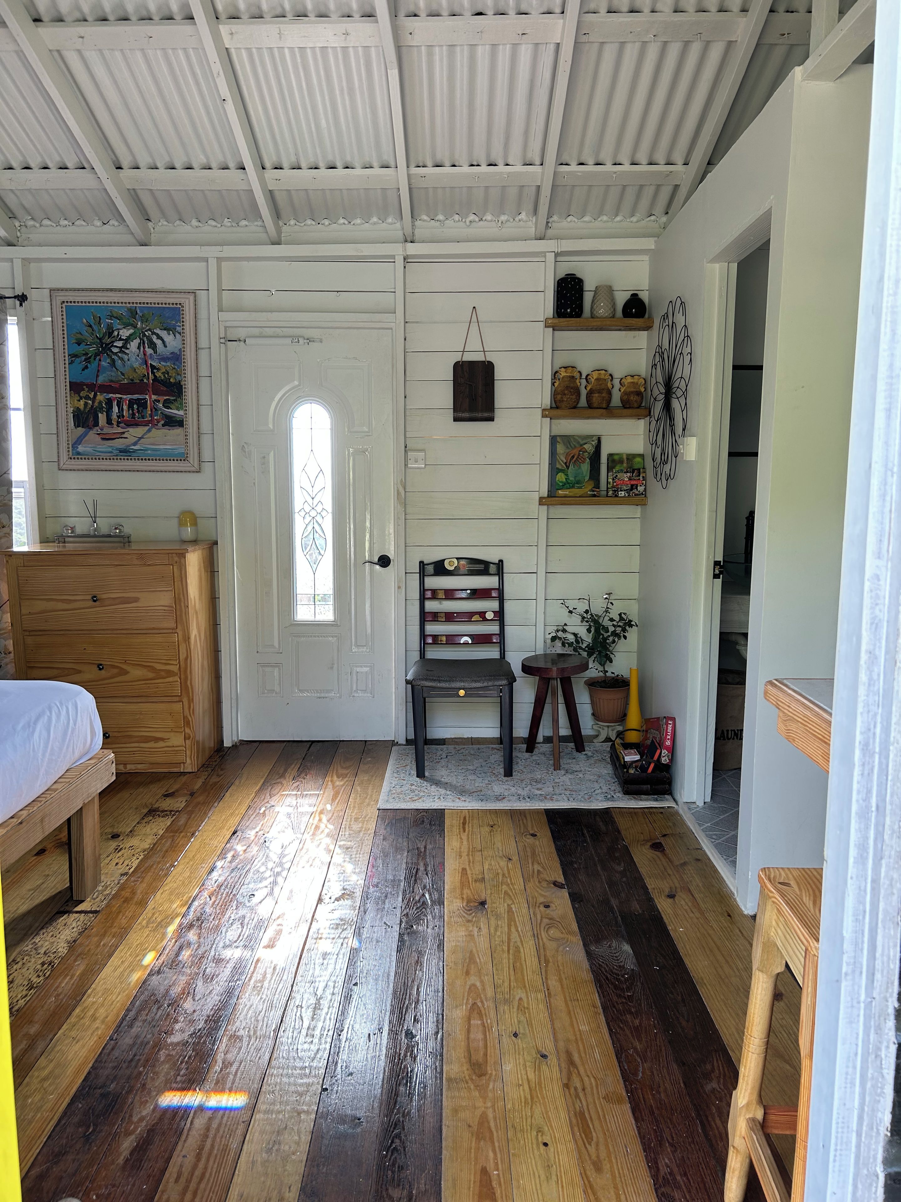 A room with a wooden floor, a bed, a chair, a shelf with decorative items, and a door with a window.