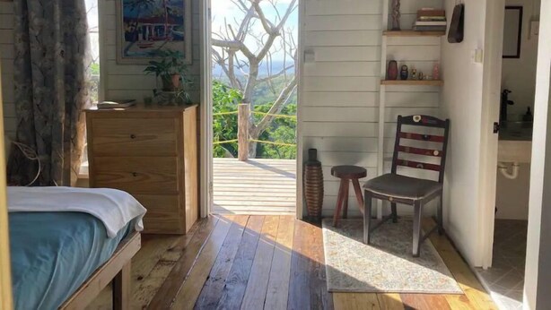 Een slaapkamer met een bed, een dressoir, een stoel, een plank met decoratieve objecten en uitzicht op groen door een geopende deur.