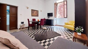 Ein Wohnzimmer mit Schachbrettmuster-Bodenbelag, einer Couch, einem gelben Sessel, einem kleinen Tisch mit Pflanze und einem Fernseher auf einem Schrank.