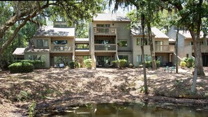 A multi-story building with balconies, a small pond, and surrounding trees. - SeaPines, Updated 2 BD Racquet Club, Exquisite View, Great Last Minute Offers (Hilton Head Island)