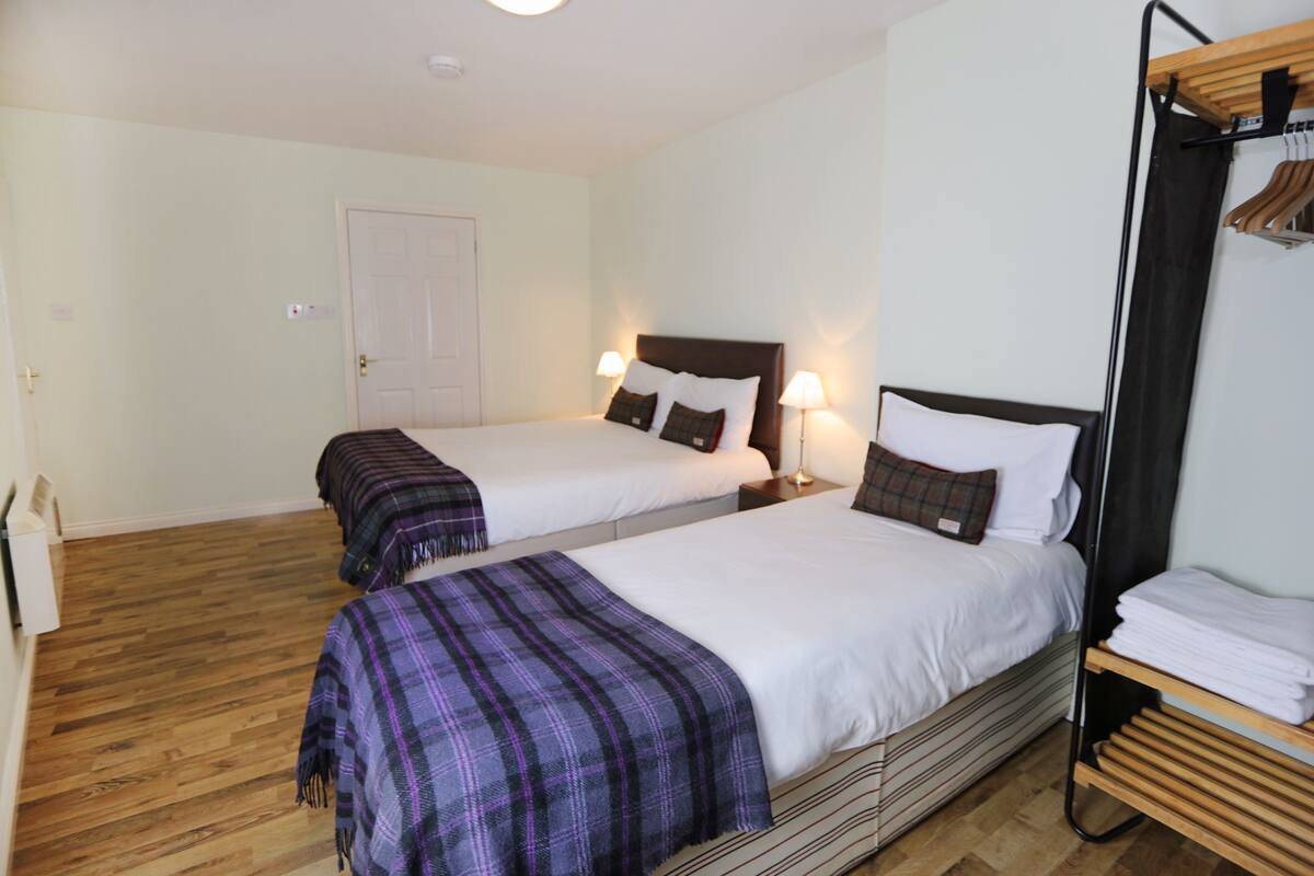 A hotel room with two beds, each with a headboard, bedside tables, and lamps. There is a wooden floor and a door in the background.