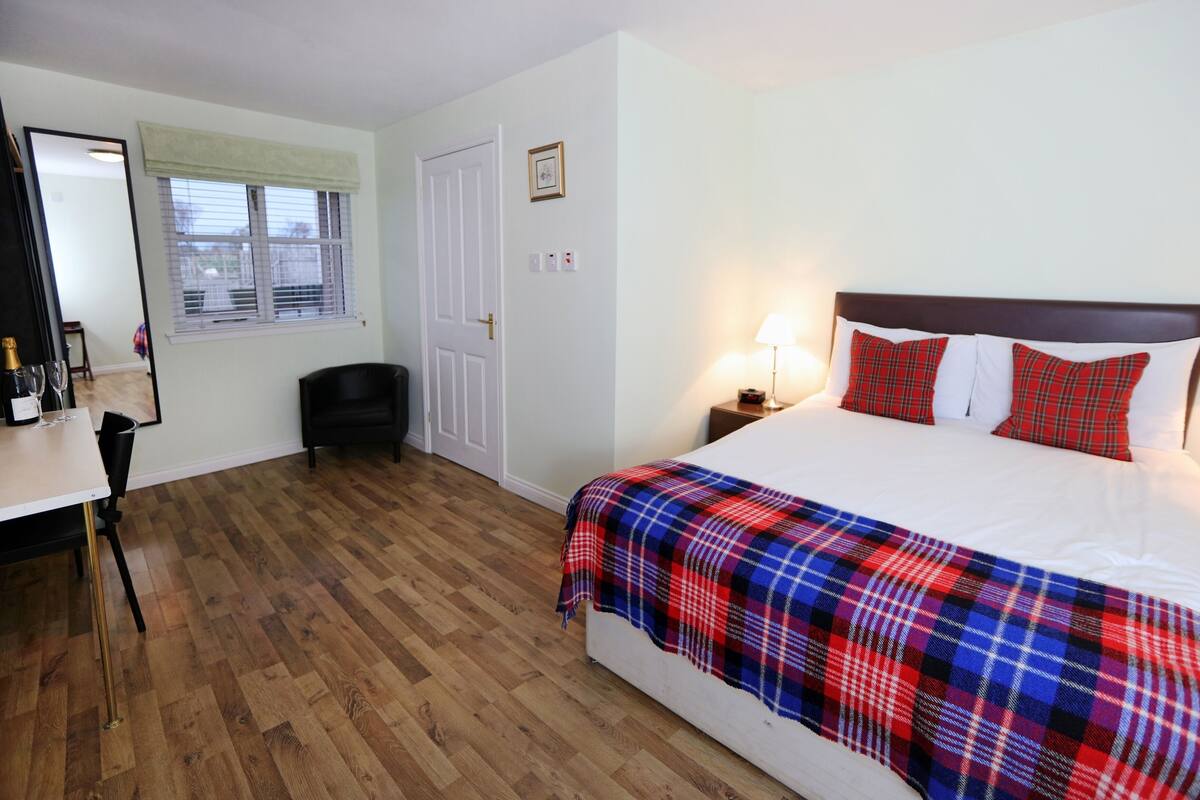 A hotel room with a bed, bedside lamps, a chair, a desk with a bottle and glasses, and a window with blinds.