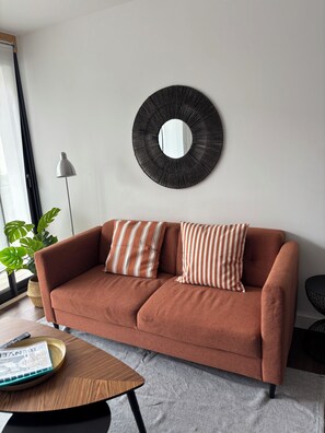 Uma sala de estar moderna com um sofá marrom, duas almofadas listradas, um espelho redondo, um abajur e um vaso com planta.