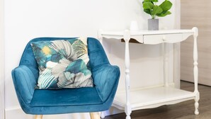 Un sillón azul verdoso con un cojín estampado con hojas, una mesita auxiliar blanca con una planta y un suelo de madera.
