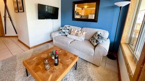 A living room with a sofa, a coffee table with a bottle and glasses, a TV, skis leaning against the wall, and a window with a lamp. - Apex Mountain ~ Mountain Tranquility (Hedley)