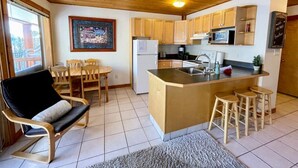 A compact kitchen with wooden cabinets, a central island, a dining area with chairs, and a black cushioned chair. - Apex Mountain ~ Mountain Tranquility (Hedley)