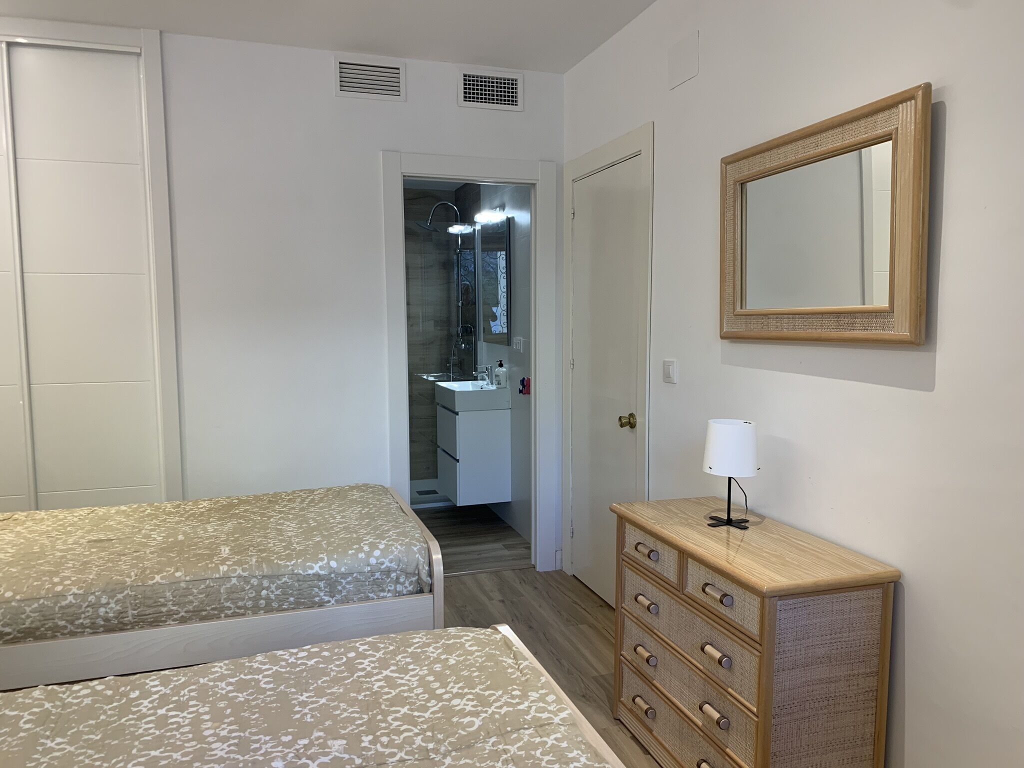 Un dormitorio con cama, cómoda, espejo y baño visible a través de una puerta abierta.