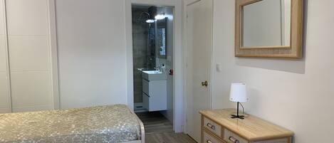 Un dormitorio con cama, cómoda, espejo y baño visible a través de una puerta abierta.
