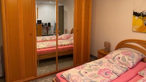 A bedroom with a wooden wardrobe, a bed with floral bedding, a bedside table, and a mirror. - Room in the countryside near Cologne/Bonn Airport (Roesrath)