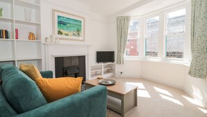 A living room with a teal sofa, a fireplace, a television, and a large window with curtains. - Bluebell Apartment (Swanage)