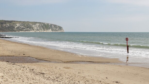 A beach with a cliff in the background, a red sign, and a wooden structure near the water. - Bluebell Apartment (Swanage)