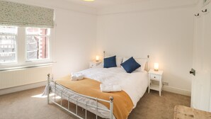 A bedroom with a bed, bedside lamps, a radiator, a window with a view of buildings, and a hanging light fixture. - Bluebell Apartment (Swanage)